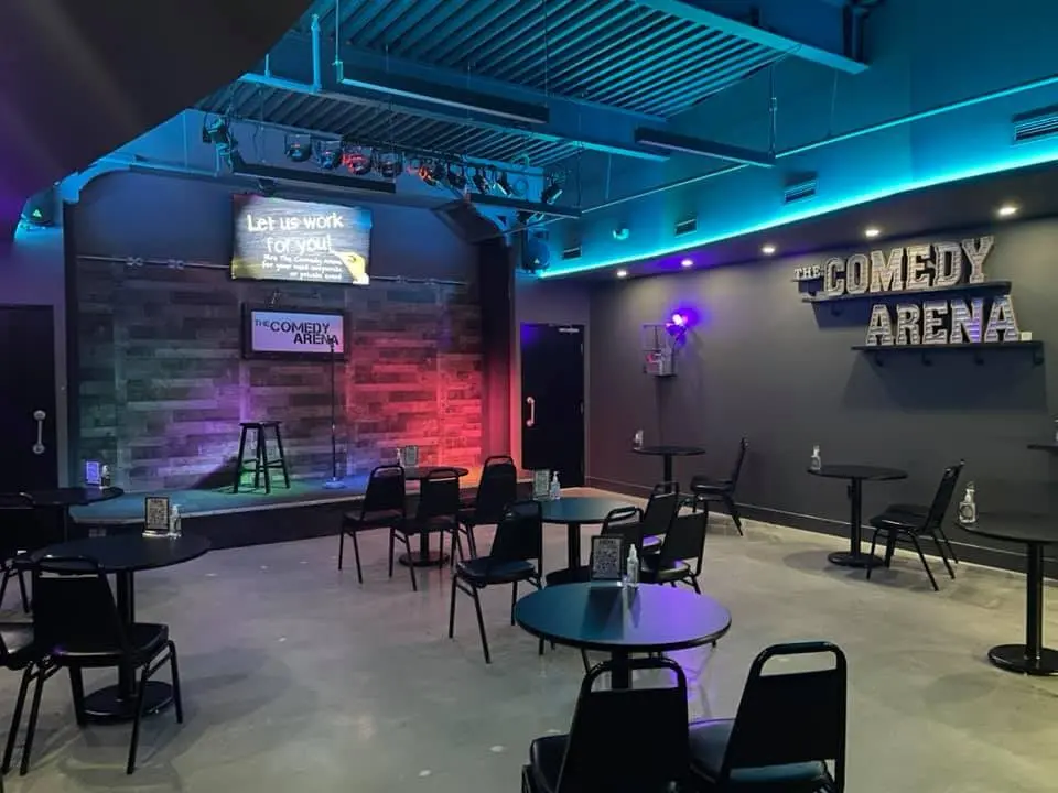 Comedy venue interior with stage, bar stools, colored lighting, and scattered seating tables and chairs