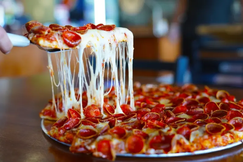 Slice of pepperoni pizza with melted cheese stretching from the slice to the pie below
