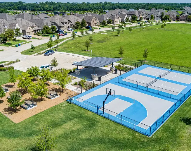 Aerial view of neighborhood park with blue pickleball courts, pavilion, seating area, and residential homes beyond green lawn