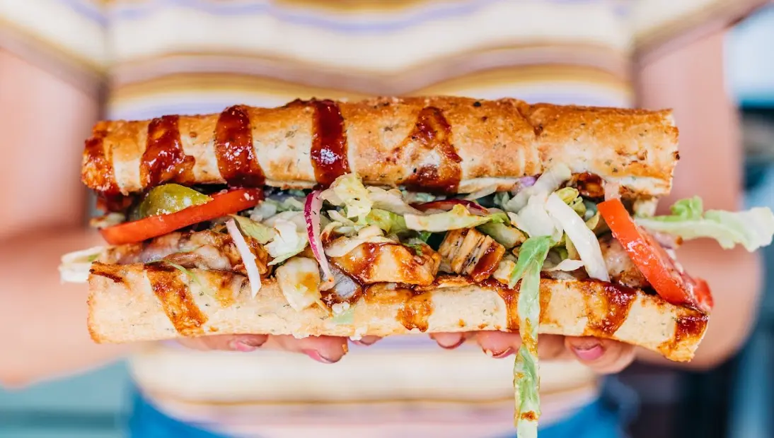 Hands holding a toasted submarine sandwich filled with meat, cheese, and shredded vegetables