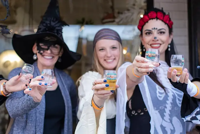 Three women in Halloween costumes holding up small beverage cups and smiling at the camera indoors