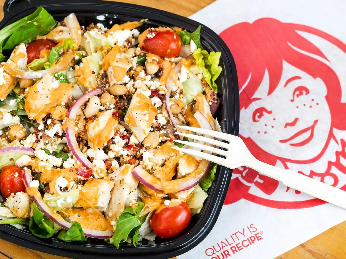 Chicken salad with crispy strips, cheese crumbles, tomatoes, red onions, and greens in a black container with a fork