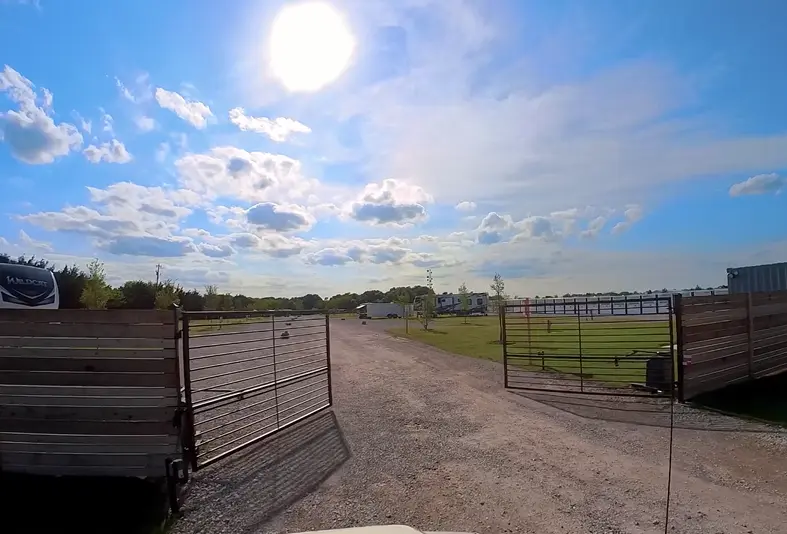 Dirt road lined with metal gates leading through an RV park with open fields and Lake Lavon visible under blue sky with sun