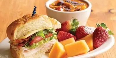 Croissant sandwich filled with lettuce and tomato, served with fresh strawberries, cheese cubes, and a bowl of soup