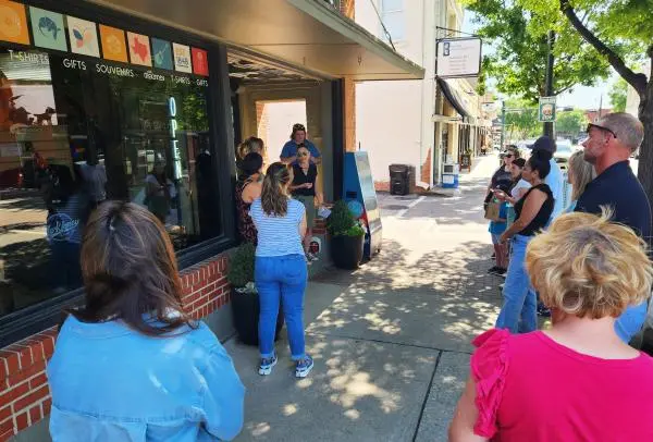Group of people gathered on a sunny tree-lined street outside a brick storefront during a walking tour