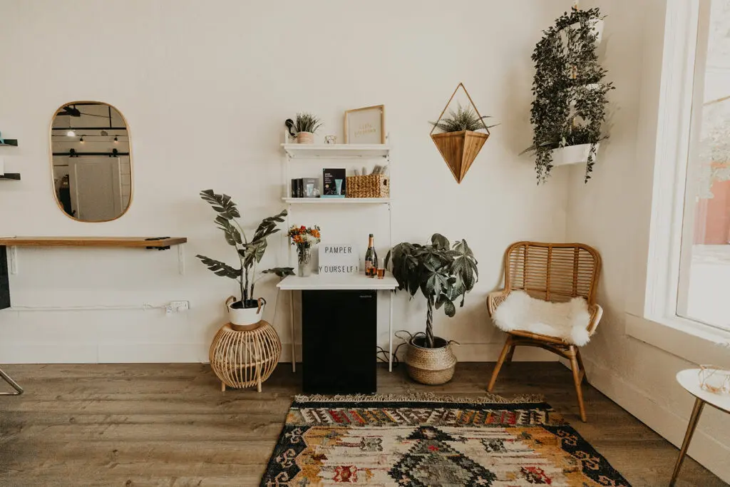 Bright studio space with wooden flooring, potted plants, woven chair, small mirror, floating shelves with decor, and geometric patterned rug