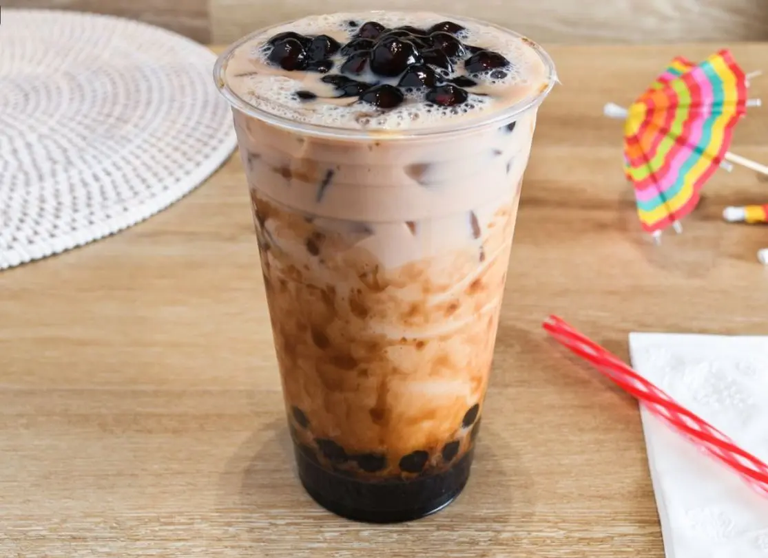 Tall glass of boba tea with tapioca pearls and creamy topping on wooden table with colorful decorations nearby
