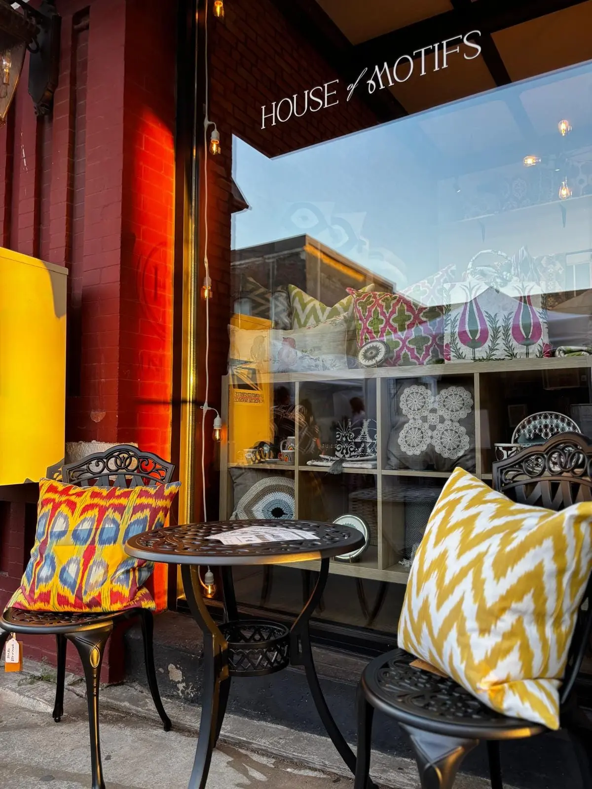 Storefront window display with ornate black chairs, patterned cushions in yellow and orange, and decorative items on shelves behind glass