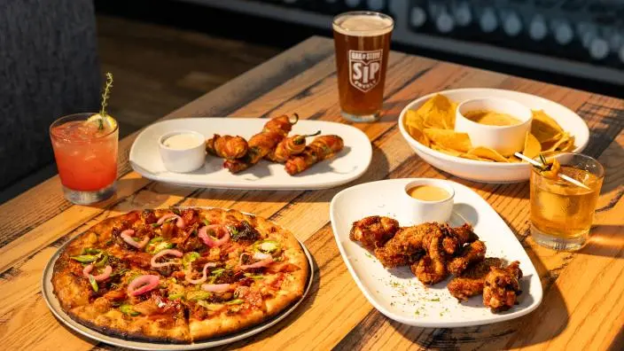 Pizza, wings, and nachos on a wooden table with beverages and dipping sauces