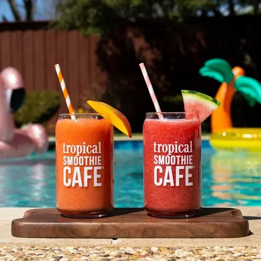 Two colorful smoothie drinks on a wooden board by a poolside, one orange with a mango slice and one pink with a watermelon slice, both labeled with the cafe