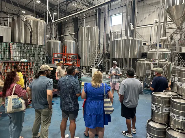 A tour group listening to the guide on the brewery floor at TUPPS, among stainless-steel tanks, kegs and pallets of cans