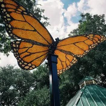 An over-sized steel Monarch butterfly sculpture with orange stained-glass-style wings, against the sky beside the green roof of a gazebo in Central Park