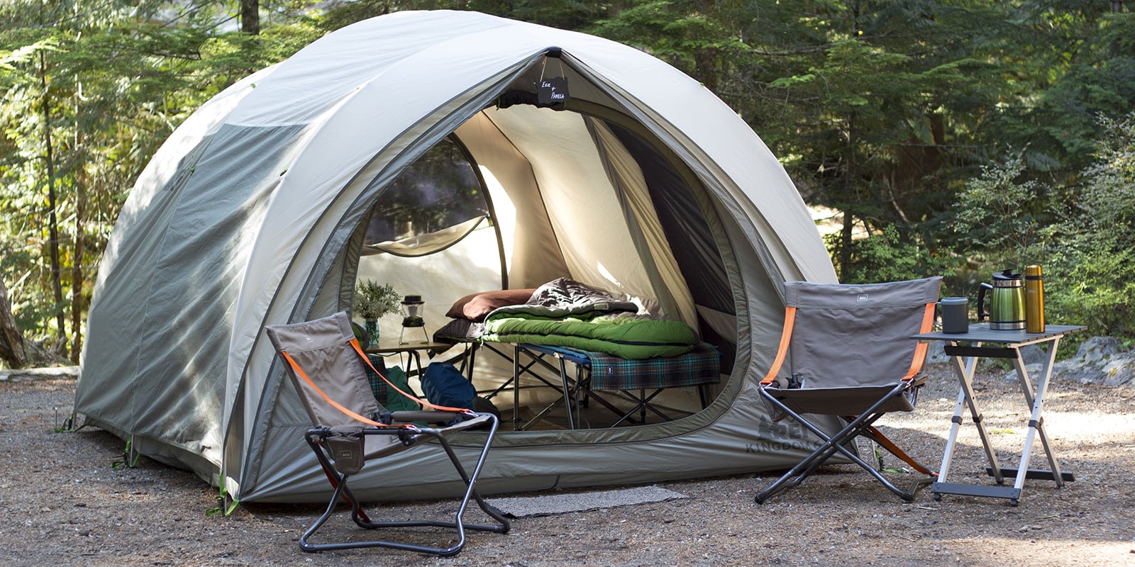 A spacious camping tent with a bed inside, chairs and a table set up in a wooded campground
