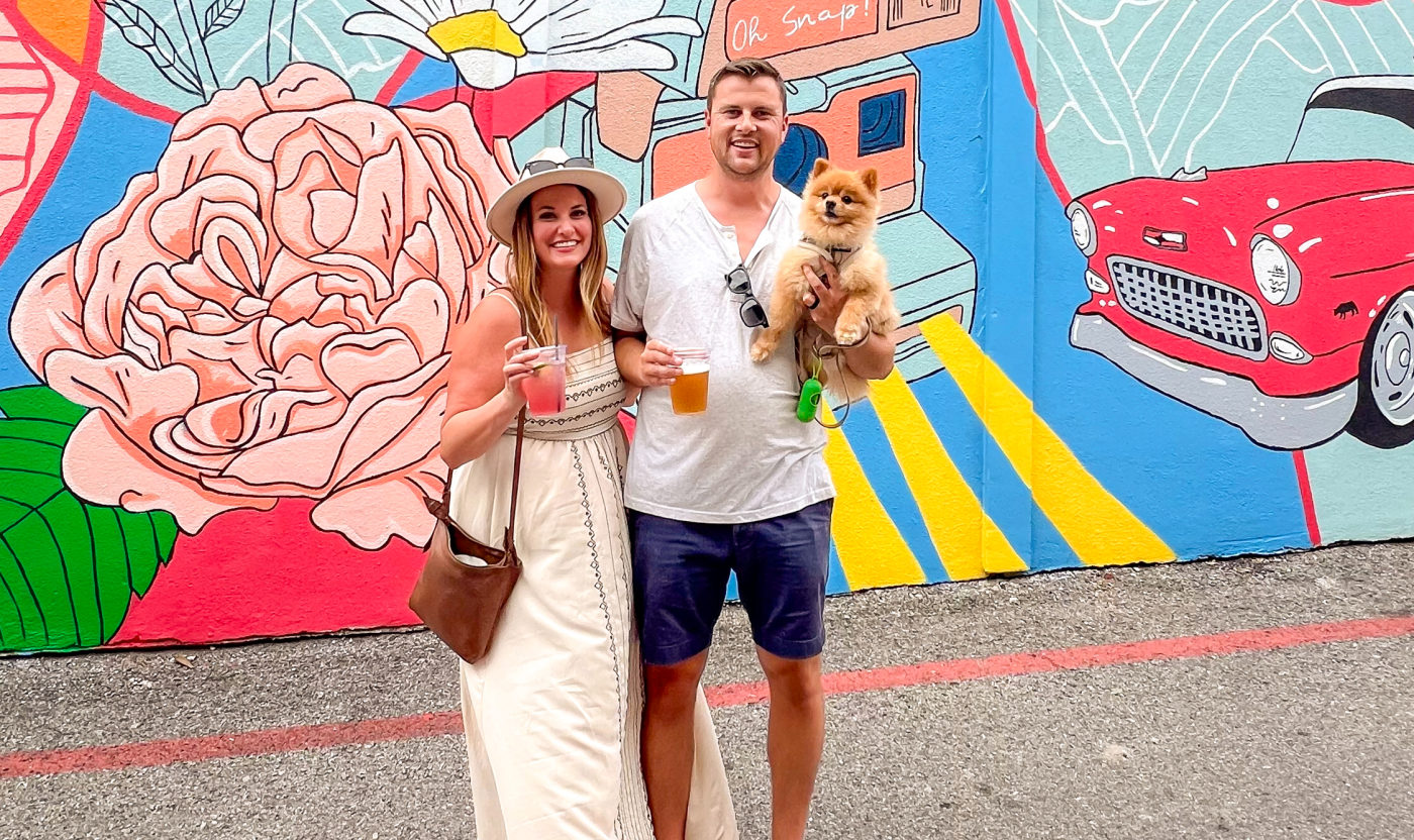 A couple standing in front of a colorful mural with cocktails and their small brown dog